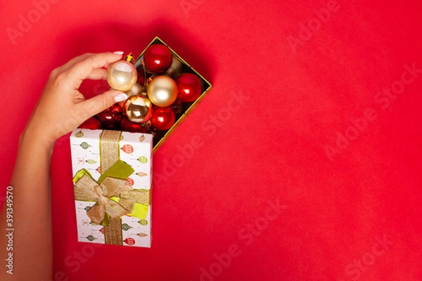 Fototapeta a woman's hand puts a ball in a green gift box with red and gold Christmas tree balls on a saturated background.