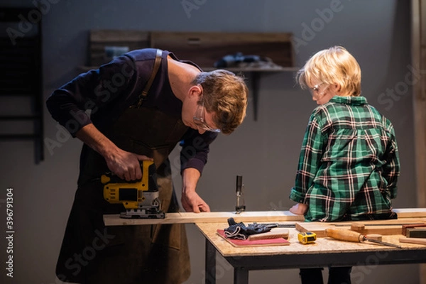 Fototapeta an experienced carpenter shows the work of various tools to his son in the workshop