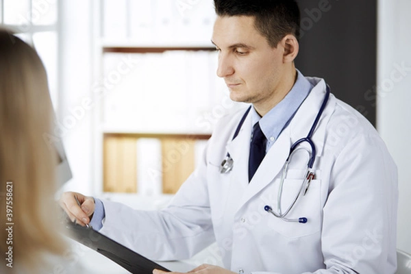 Fototapeta Friendly male doctor and patient woman discussing current health examination while sitting in sunny clinic. Perfect medical service in hospital