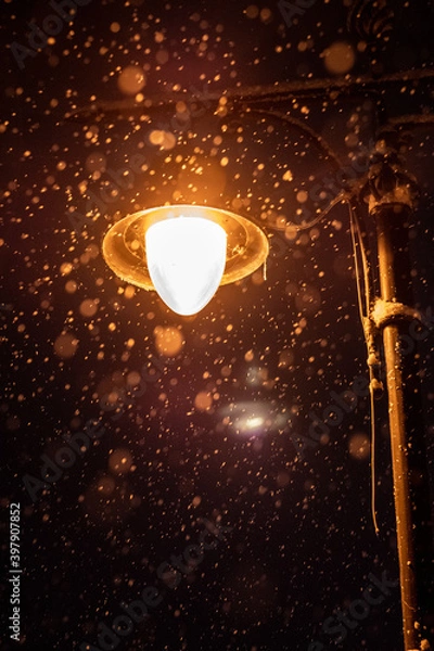 Fototapeta Detail of a street lamppost at night with close up on the snow falling. Selective focus.