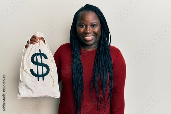 Fototapeta Young black woman with braids holding dollars bag looking positive and happy standing and smiling with a confident smile showing teeth