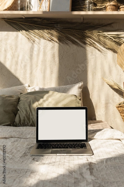 Fototapeta Blank screen laptop. Warm sunlight shadows on the wall. Parisian vibes. Warm bohemian style home interior design. Copy space mockup template. Hygge bedroom with bed, pillows, linens, decorations.