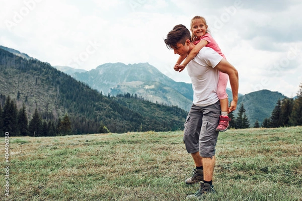 Fototapeta Young man holding little girl on his back. Child playing with her elder brother riding on his back enjoying summer day together. Happy siblings playing in the field during vacation trip in mountains