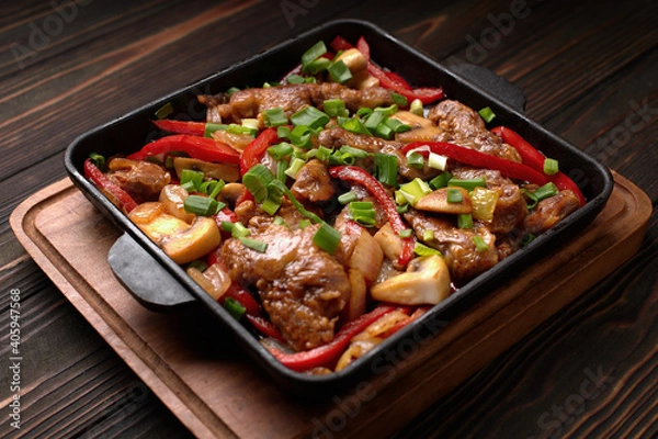 Fototapeta Fried meat with vegetables, green onions, bell peppers and mushrooms, in a frying pan, on a wooden background