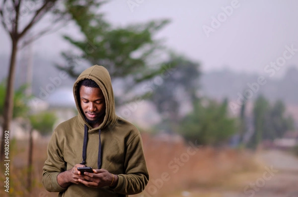 Fototapeta young african man standing outside and delighted while using his smart phone