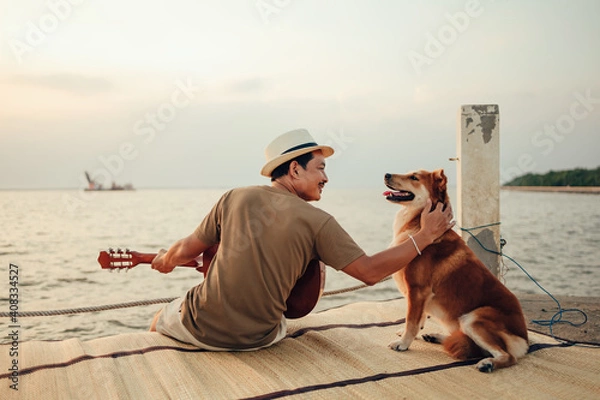 Fototapeta A man wear straw hat and playing guitar music song near the sea sunset and stroking the head dog pet.