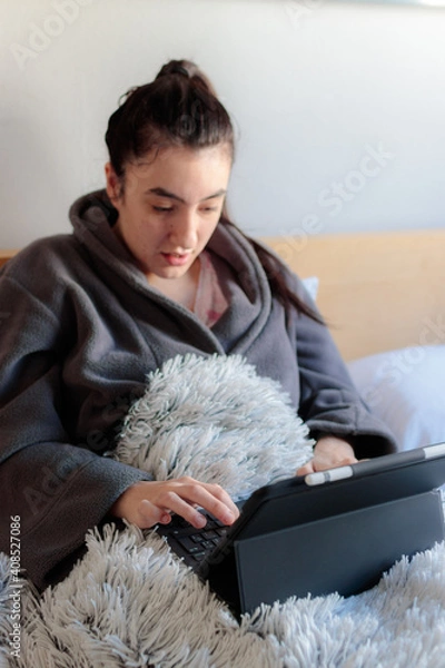 Fototapeta Mujer caucásica joven sentada en la cama de su habitación con ropa e pijama tapada con una manta usando una tablet estudiando en casa. 