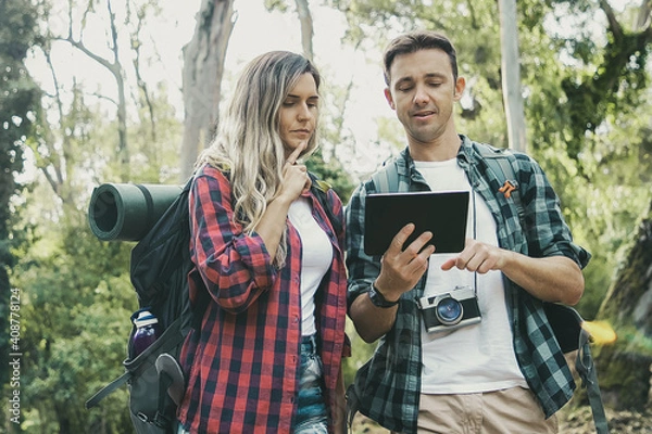 Fototapeta Caucasian couple checking road on map via tablet. Young couple of travelers trekking in forest together with backpacks. Green trees on background. Tourism, adventure and summer vacation concept