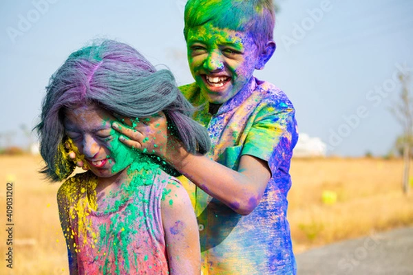 Fototapeta Young kid from the back applying holi colors to girls face during holi festival celebration - Concept of kids having fun by playing holi during festive.