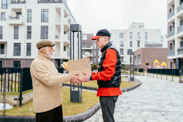 Fototapeta Profile shot of an elderly senior man taking to a food delivery male with a grocery box outdoor with building on background. Meal basket as social help and support.