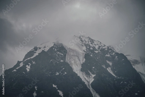 Fototapeta Dark atmospheric mountain landscape with glacier on black rocks in lead gray cloudy sky. Snowy mountains in low clouds in rainy weather. Gloomy landscape with black rocky mountains with snow in fog.