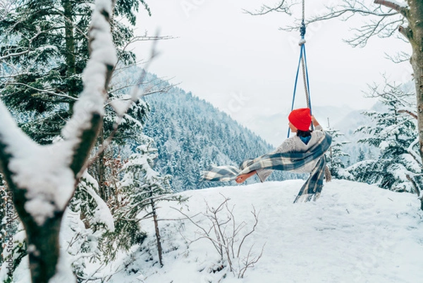 Fototapeta A young woman in warm clothes, checkered scarf and Red Cap swinging on a swing between forest trees with picturesque snowy mountain view. Wintertime vacation concept image.