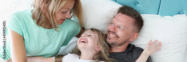 Fototapeta Happy family lies in bed. Girl lies on her dad and laugh. Woman look at her daughter and is surprised. Cheerful man look at his wife.