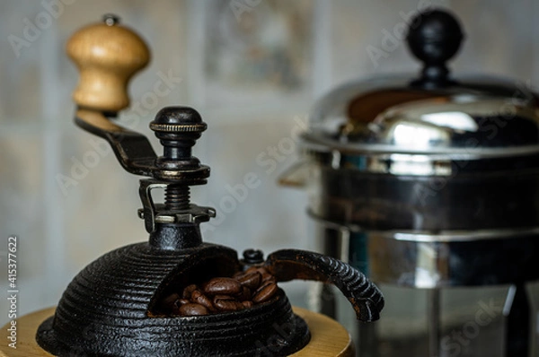 Obraz Coffee beans in a manual grinder with a French press in the background.