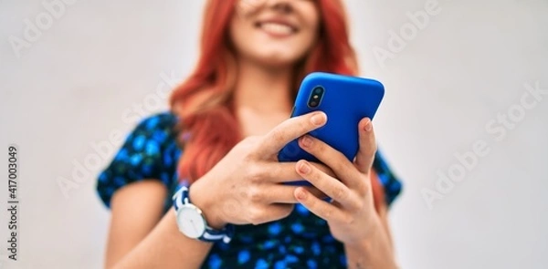 Fototapeta Young redhead girl smiling happy using smartphone at the city.