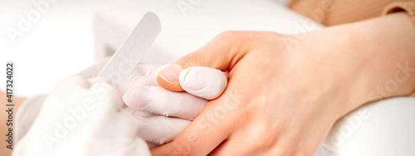 Fototapeta Manicure master wears white gloves with nail file doing manicure on female fingernails in a nail salon