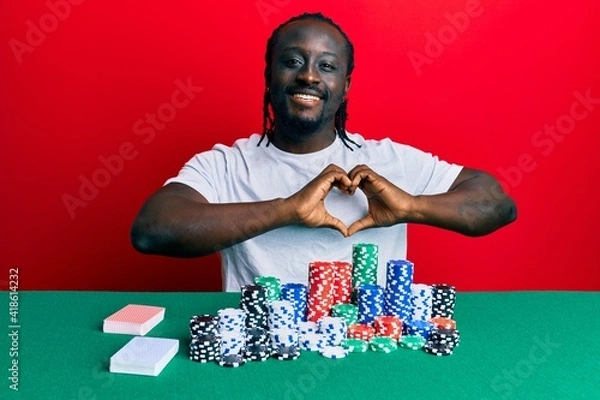 Fototapeta Handsome young black man sitting on the table with poker chips and cards smiling in love showing heart symbol and shape with hands. romantic concept.