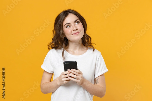 Fototapeta Young happy dreamful wistful pensive student woman 20s wear white t-shirt holding mobile cell phone chatting browsing internet look aside isolated on yellow orange color background studio portrait.