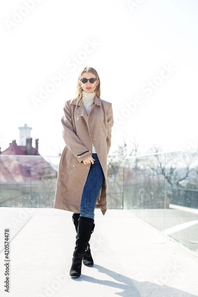 Fototapeta Young girl wearing sunglasses with long blonde hair in the wind, wearing a long coat outside