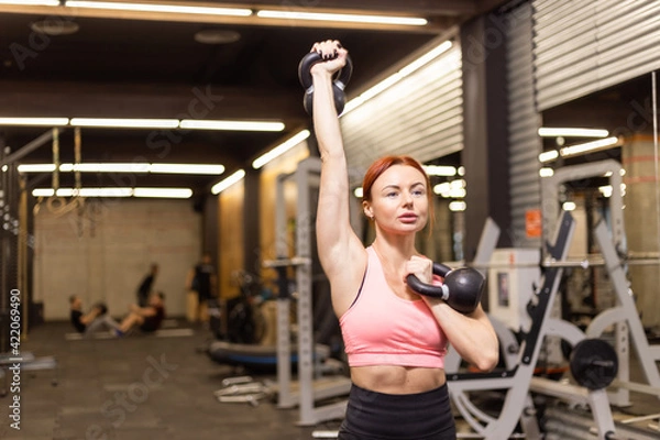 Fototapeta Intensive workout red-haired woman with kettlebells in her hand in modern gym