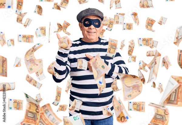 Fototapeta Senior handsome man wearing burglar mask and t-shirt pointing to the back behind with hand and thumbs up, smiling confident