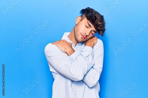 Fototapeta Young hispanic man wearing casual clothes hugging oneself happy and positive, smiling confident. self love and self care