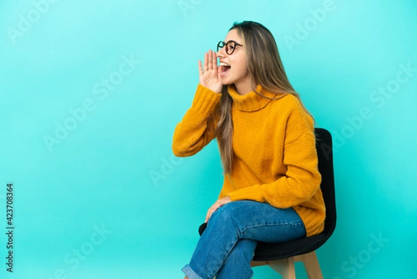 Fototapeta Young caucasian woman sitting on a chair isolated on blue background shouting with mouth wide open to the lateral