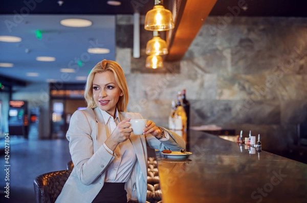 Fototapeta A beautiful middle-aged businesswoman sitting in a hotel cafe and enjoying her coffee. She waiting for a business meeting. Free time, enjoyment, respite, lifestyle