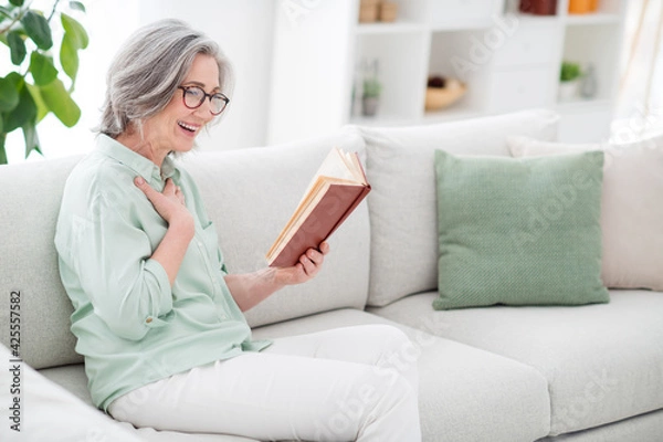 Fototapeta Photo of cheerful person sit on couch reading book arm on chest laughing have fun free time weekend indoors