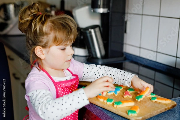 Fototapeta Cute little toddler girl and fresh baked homemade Easter or spring cookies at home indoors. Adorable blond child with apron with bunny and carrot cookie in domestic kitchen. Child eating cookie