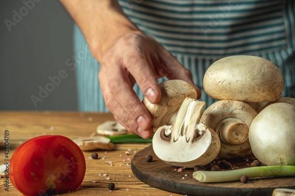 Fototapeta Chef is holding a mushroom in his hand over a wooden table with vegetables and other ingredients for cooking the dish