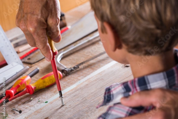 Fototapeta Grandfather teaches boy to work on bench with joinery tools