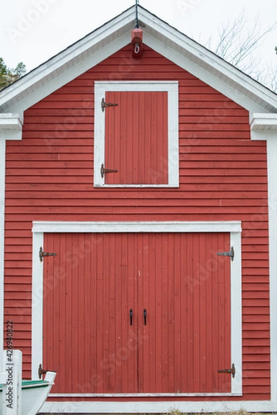 Fototapeta An old two storey red barn with large double red doors, white trim, and a small closed wood loft window. The sky in the background is grey. There's a wooden boat on the ground in front of the doors. 