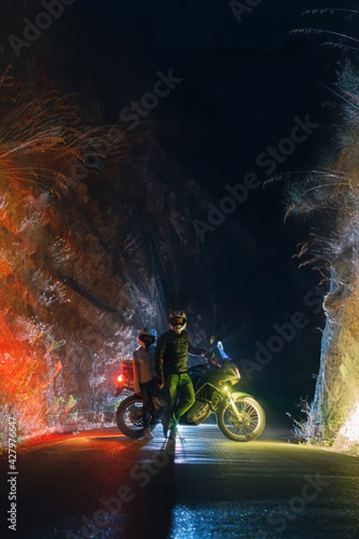 Fototapeta Motorcyclist biker man with a motorcycle for long journeys. A road in a narrow canyon. Night and lights. Tourism and vacation. Vertical photo.