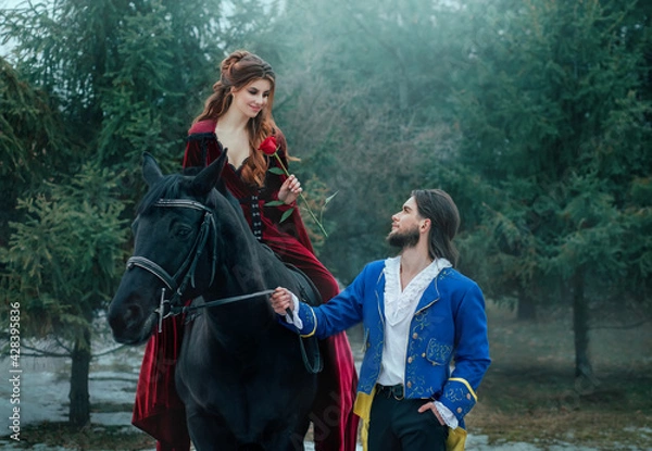 Fototapeta Medieval couple in love. man meets woman and gives rose in winter forest. Vintage clothing red long dress. Blue costume tailcoat caftan. Prince and princess together. Black horse. redhead hairstyle.