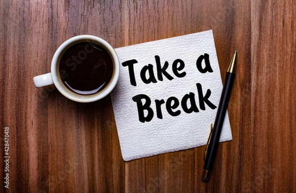 Fototapeta On a wooden table next to a white cup of coffee and a pen is a white paper napkin with the words TAKE A BREAK