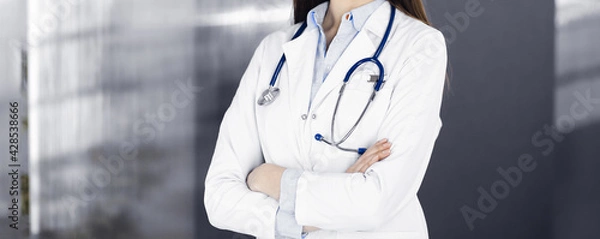 Fototapeta Unknown young woman doctor is standing with arms crossed in clinic. Medicine concept during Coronavirus pandemic