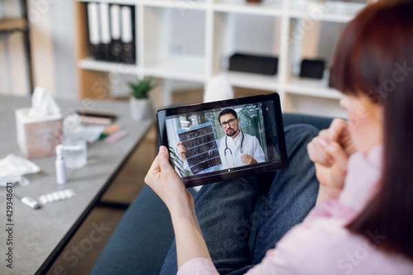 Obraz Tablet monitor view over woman shoulder. Sick woman with cough, listens to male therapist. Doctor gives recommendations for home treatment of his patient. Online medical service app