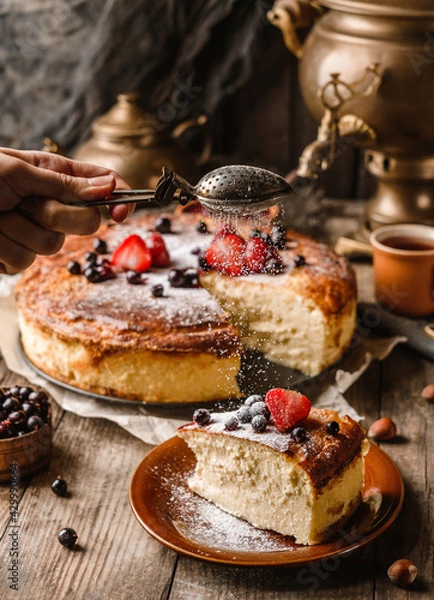 Fototapeta Woman hand sprinkles icing sugar on a cheesecake with berries on rustic wooden table with samovar and teapot, complex composition. Sweet dessert, close up