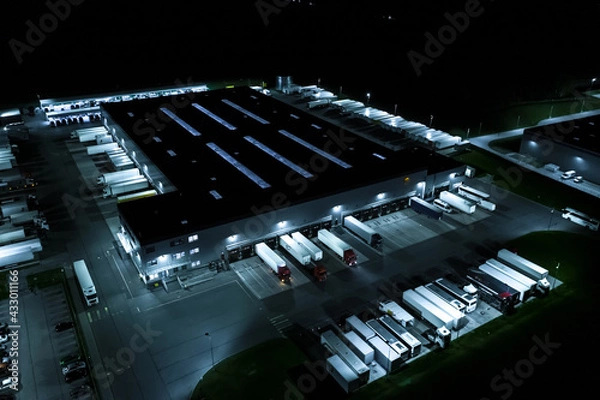 Fototapeta Aerial view of goods warehouse at Night. Logistics center in industrial city zone from above. Aerial view of trucks loading at logistic center. View from drone.