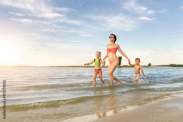 Fototapeta Young adult attractive slim sporty mother enjoy having fun running water by lake or sea sand breach with two cute little siblings against blue sky on summer day. Summertime family vacation concept