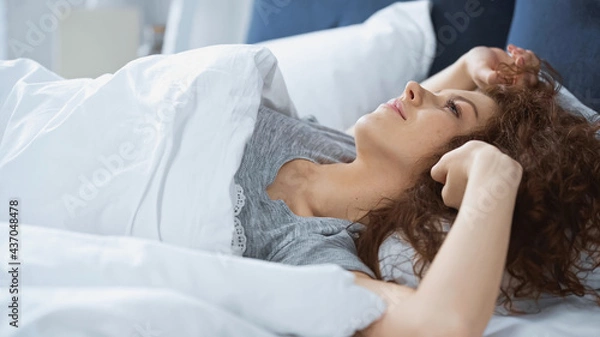 Fototapeta pleased young and curly woman lying on bed in morning