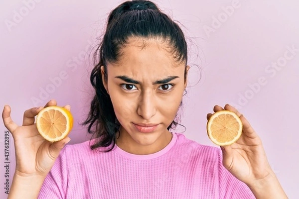 Fototapeta Hispanic teenager girl with dental braces holding lemon skeptic and nervous, frowning upset because of problem. negative person.