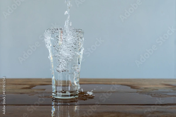 Fototapeta Water flows into a glass placed on a wooden bar. Grey background