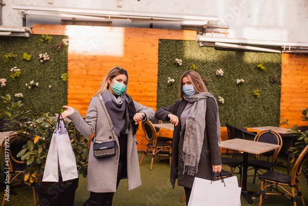 Fototapeta Two beautiful young blonde women with medical face masks greet each other with their elbows in the city. Shopping during the COVID-19 coronavirus epidemic. Social distance