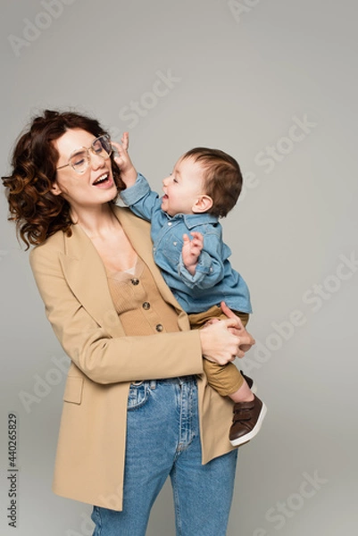 Fototapeta cheerful toddler boy reaching glasses on curly mother smiling isolated on grey