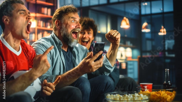 Fototapeta Night at Home: Three Soccer Fans Sitting on a Couch Watch Game on TV, Use Smartphone App to Online Bet, Celebrate Victory when Sports Team Wins. Friends Cheer Eat Snacks, Watch Football Play.