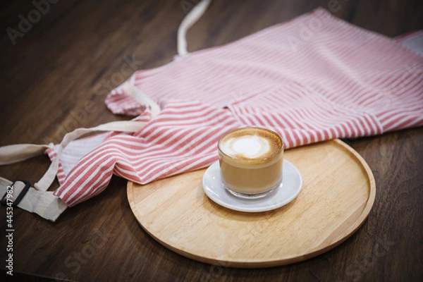 Fototapeta Cappuccino in a clear glass and served with an apron near the brown table.