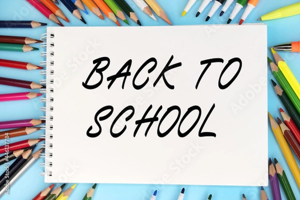 Fototapeta Back to school is written on a notebook that lies on a blue background among colored pencils and markers. Educational concept