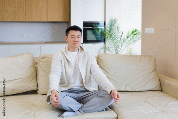 Fototapeta young asian man sits on sofa in lotus position and meditates in don